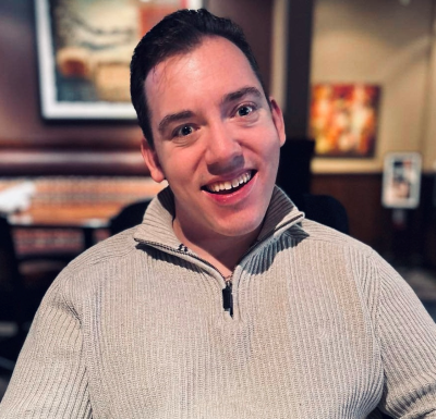 Keenan is seated at a table, wearing a light a gray sweater. He has dark brown hair, grey-green eyes and an open inviting smile.